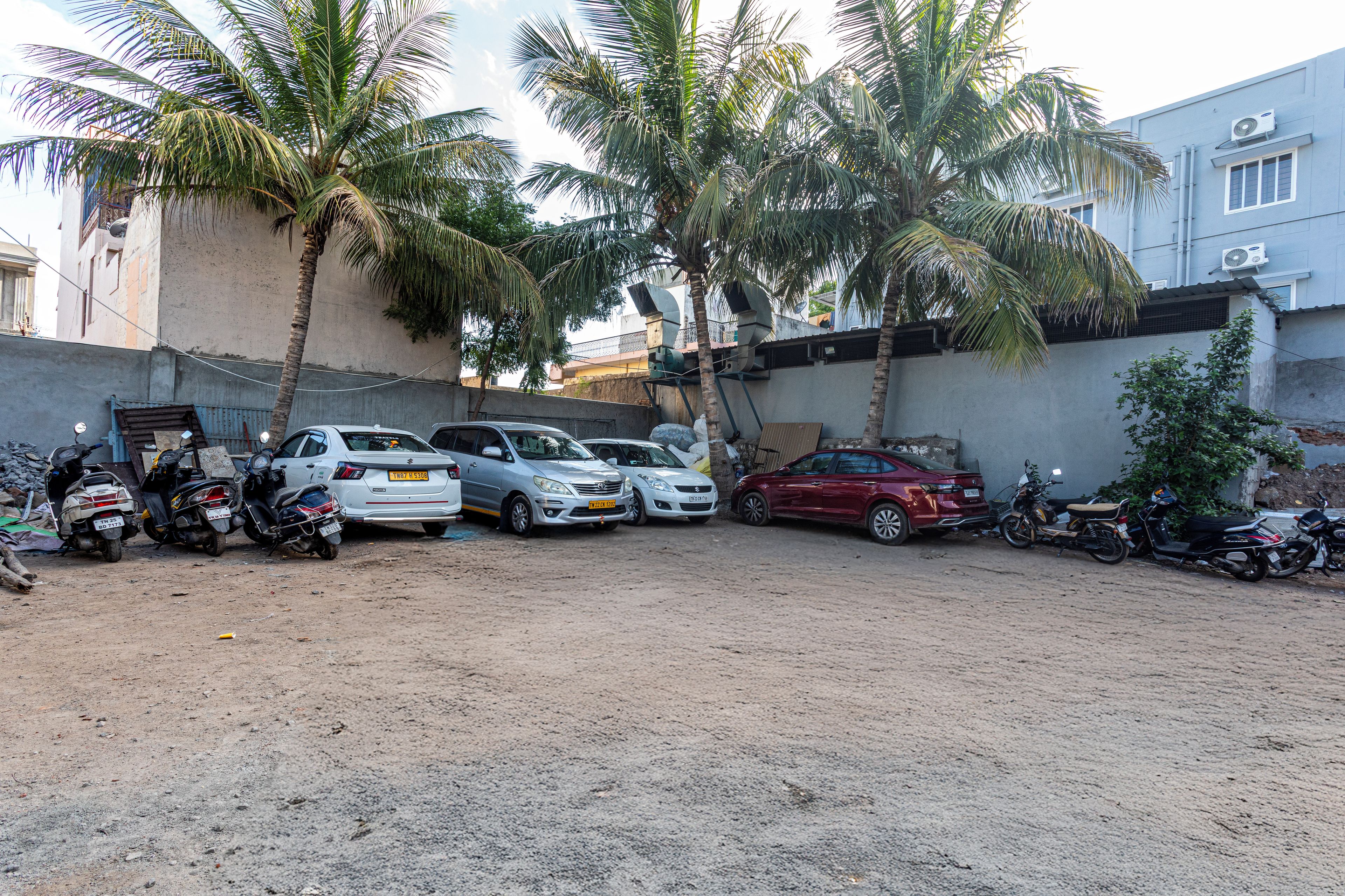 A parking area with multiple cars and motorcycles, surrounded by a concrete wall and palm trees.