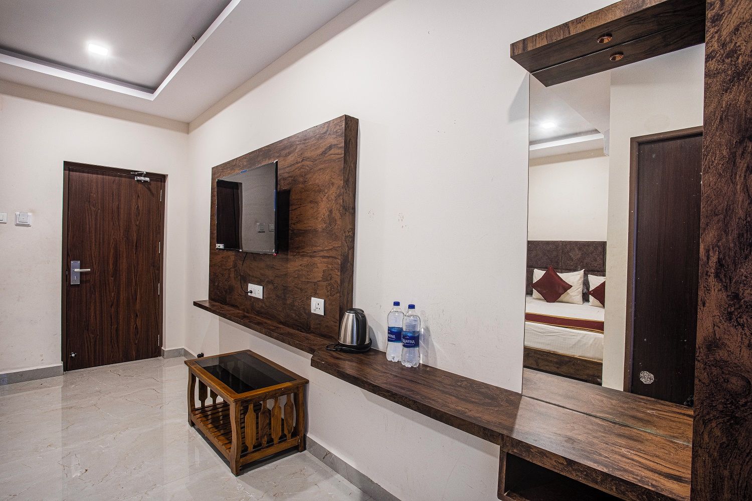 Une chambre d’hôtel avec une coiffeuse en bois, un miroir, un lit agrémenté d’oreillers à motifs et une petite table en bois sur laquelle se trouvent une bouilloire et des bouteilles d’eau.