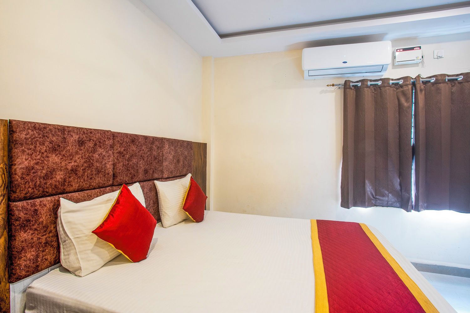 A hotel room with a bed, a headboard, a window with curtains, and an air conditioning unit.