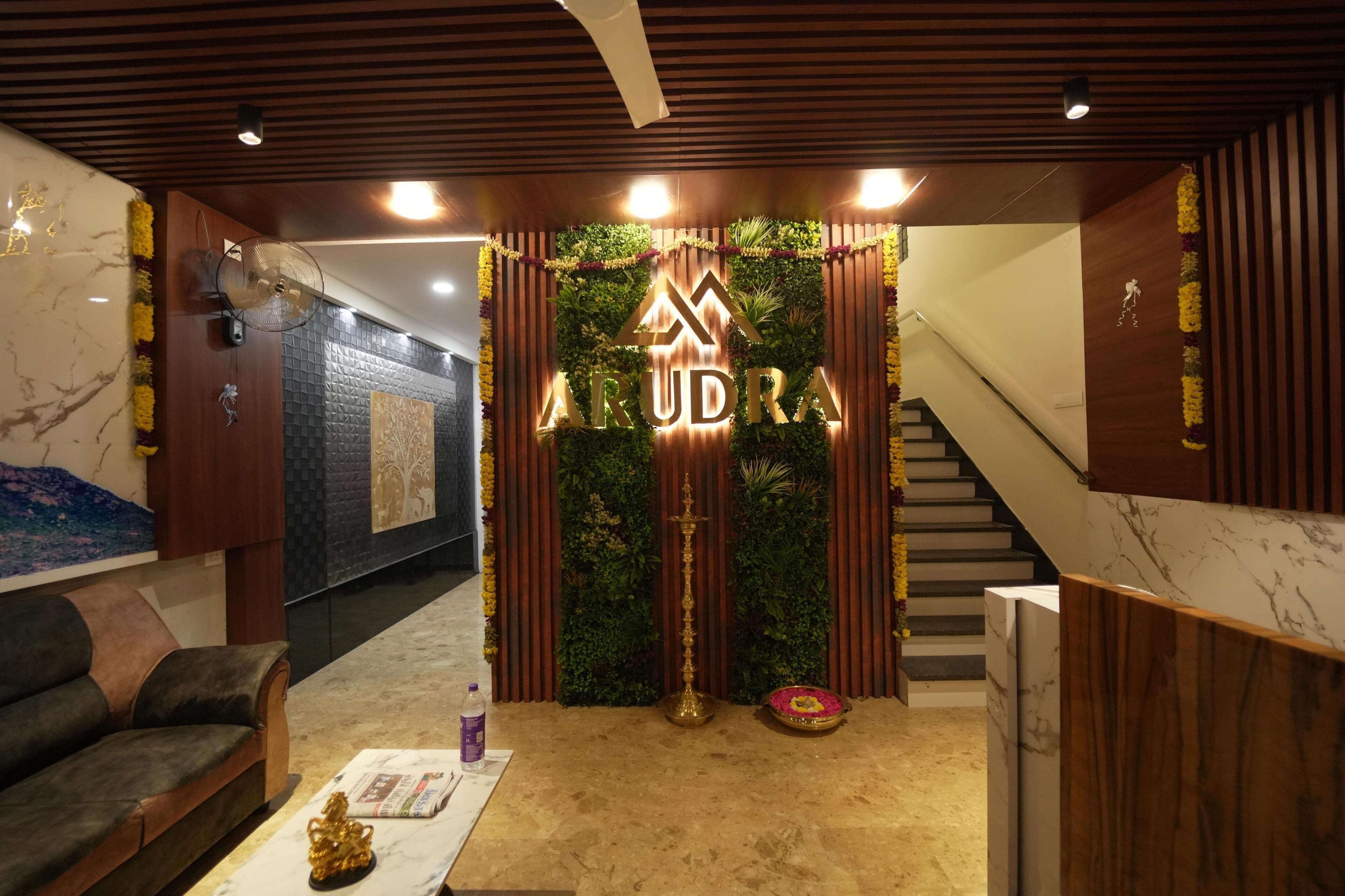 A reception area with a wooden reception desk, a sofa, a fan, and a decorative wall with the name 'ARUDRA'.
