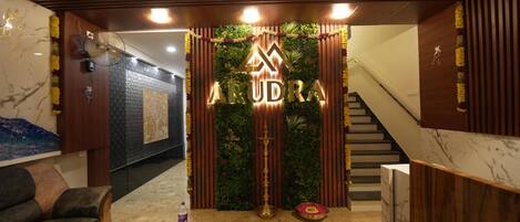 A reception area with a wooden reception desk, a sofa, a fan, and a decorative wall with the name 'ARUDRA'.