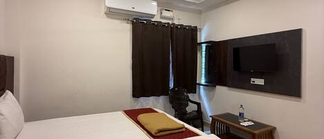 A hotel room with a bed, a ceiling fan, an air conditioning unit, a television mounted on the wall, a small table with a bottle, and a chair.