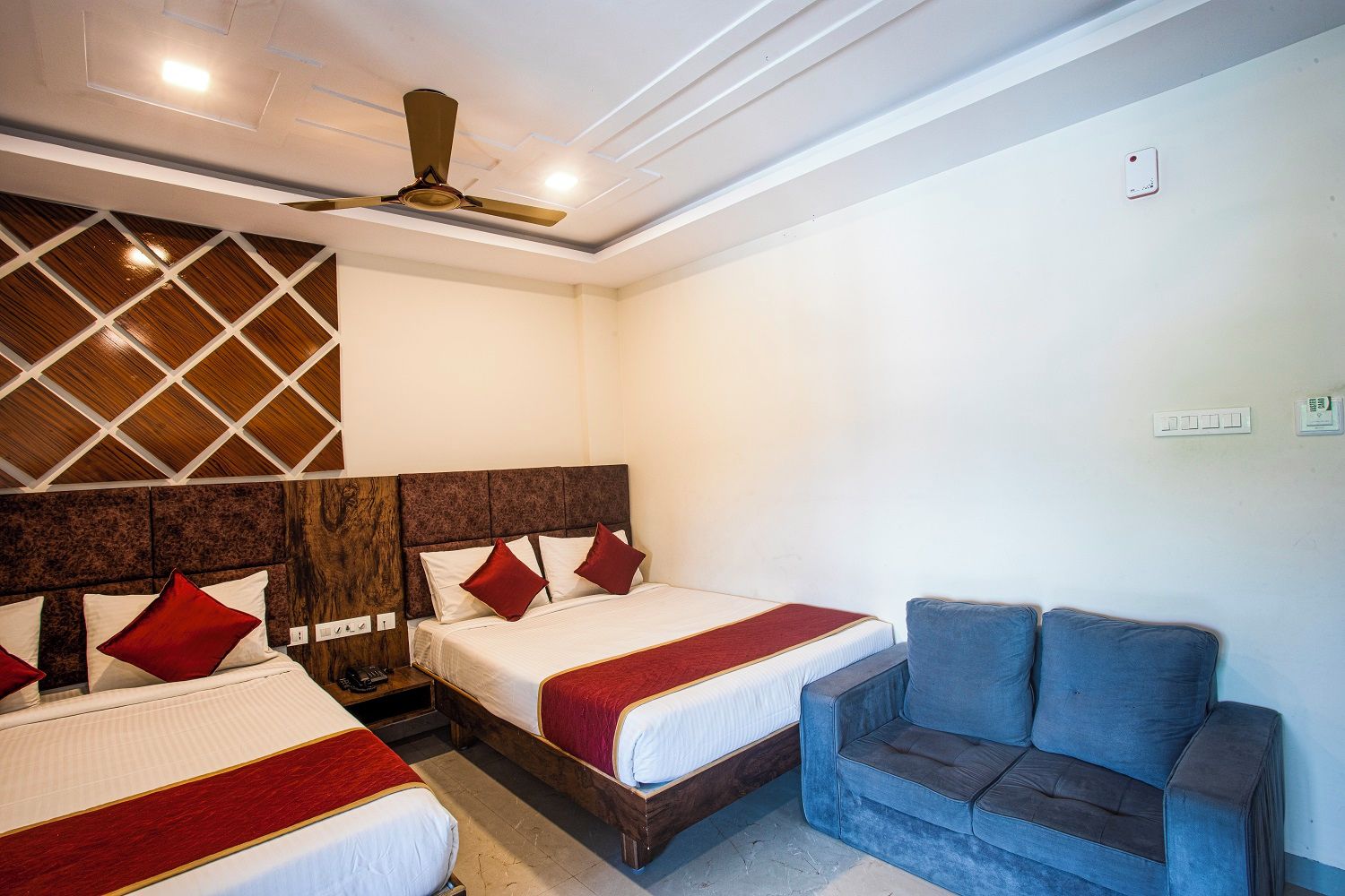 A hotel room with two beds, a sofa, a ceiling fan, and a wall-mounted light fixture.