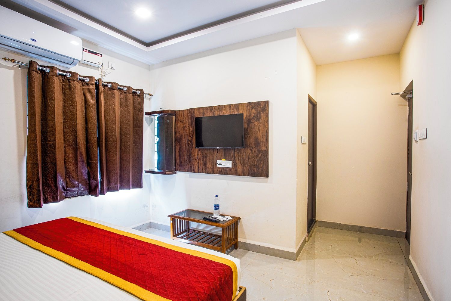 A hotel room with a bed, a TV mounted on the wall, a small table, and a door leading to another room.
