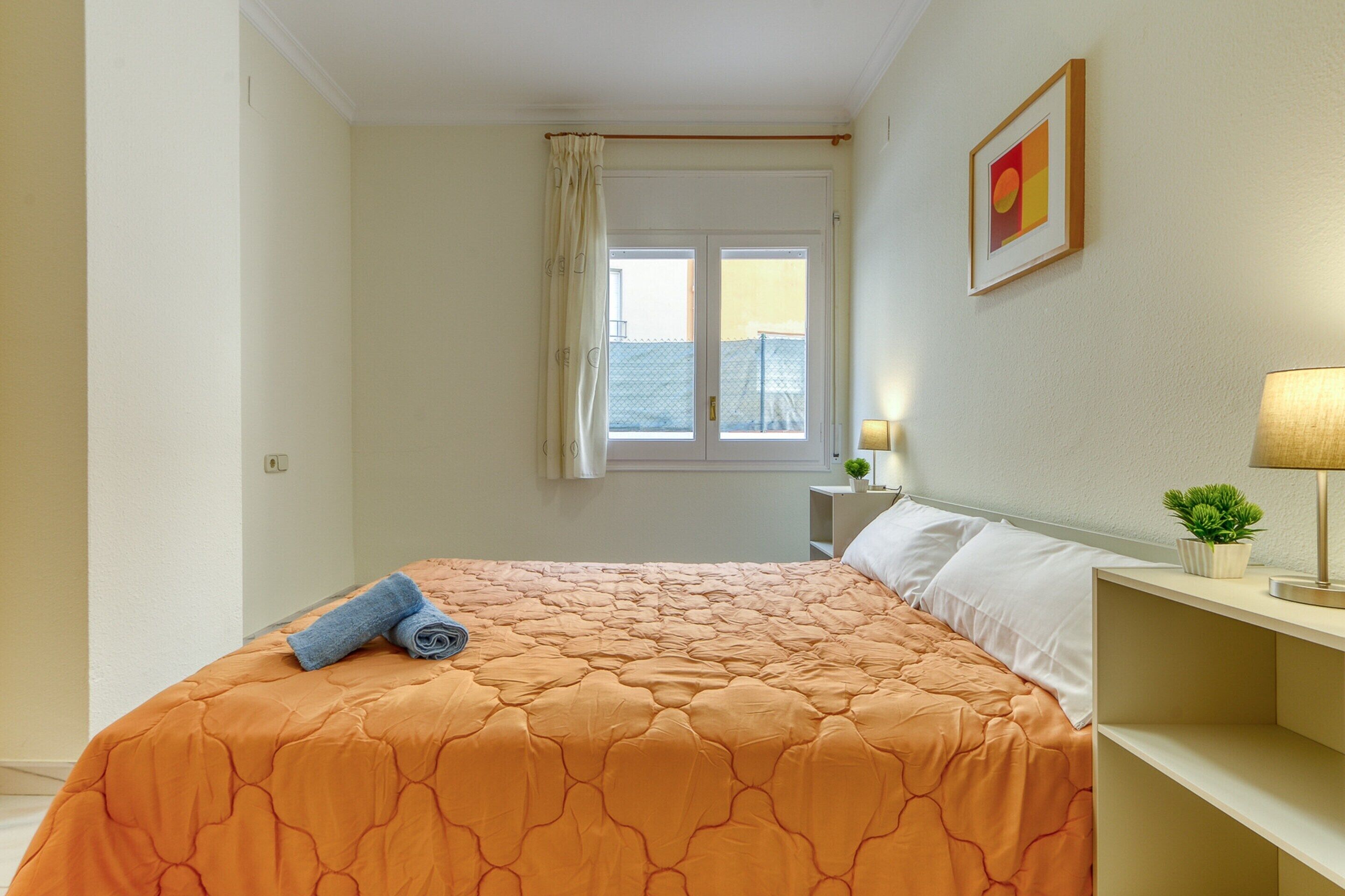 A neatly made bed with an orange bedspread, a small bedside table with a lamp, and a window with curtains.