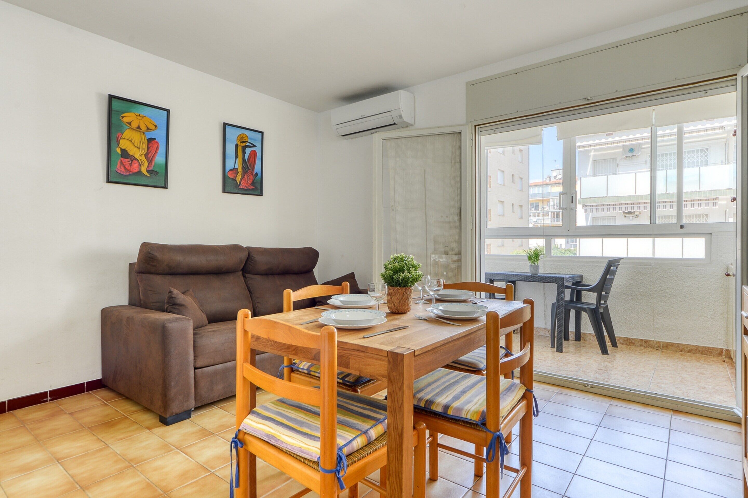 A living room with a sofa, dining table, and chairs. There are two framed artworks on the wall, and an air conditioning unit above the sofa.