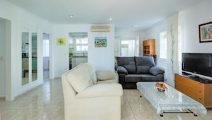 A spacious living area with a sofa, a coffee table, and a television. There is a kitchen in the background with cabinets and a sink. - Tranquil Holiday Home in Empuriabrava (Castello d'Empuries)