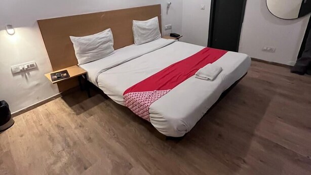 A modern hotel room with a wooden floor, a bed with white bedding and a red accent, a nightstand, and a wall-mounted mirror. - Hotel Arena  (Gurugram)