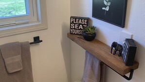 A bathroom with a toilet, a towel rack, a window, a sign saying "PLEASE SEAT YOURSELF", a shelf with a plant and a decorative item, and a framed picture on the wall. - Boho Bungalow  2-bedroom house Professionally Cleaned in delightful Fort Smith (Fort Smith)