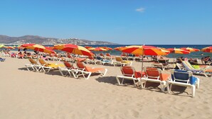A beach with rows of sun loungers and umbrellas, a clear sky, and a distant view of a town. - Apartment Empuriabrava Near Sandy Beach (Castello d'Empuries)