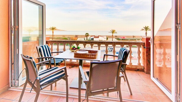 A balcony with a table and chairs, overlooking a beach with palm trees and a clear sky. - Apartment Empuriabrava Near Sandy Beach (Castello d'Empuries)