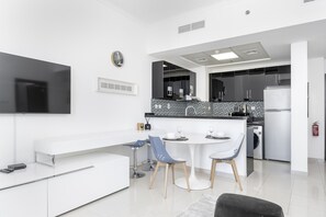Una cocina moderna con armarios blancos, un protector contra salpicaduras negro y un horno empotrado. Hay una zona de comedor con una mesa blanca y sillas azules. Un televisor de pantalla plana está montado en la pared sobre la cocina.