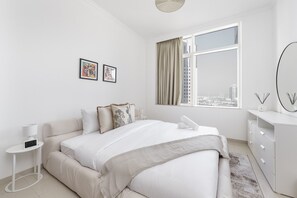 A modern bedroom with a large bed, a side table, a dresser, and a window with a city view.