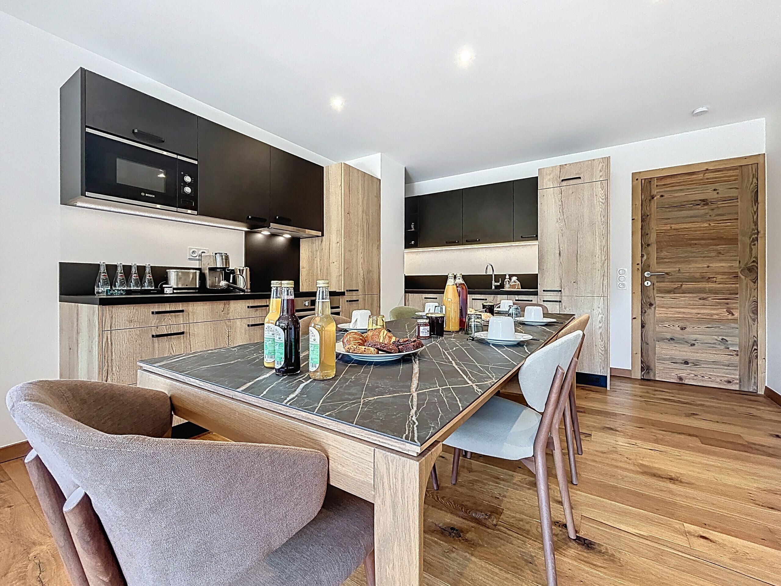 A modern kitchen with dark cabinets, a black microwave, and a dining table set with chairs. There are bottles on the table, and a wooden door is visible in the background.