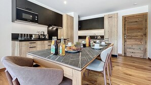 A modern kitchen with dark cabinets, a black microwave, and a dining table set with chairs. There are bottles on the table, and a wooden door is visible in the background. - Tamaris - Morzine - secteur Planet (Morzine)