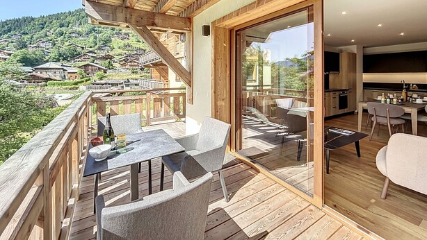 A wooden deck with a table and chairs, a glass door leading to a kitchen, and a view of a hillside village. - Tamaris - Morzine - secteur Planet (Morzine)