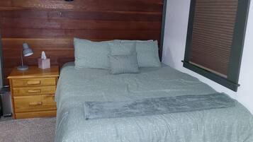 Une chambre à coucher avec une tête de lit en bois, un lit recouvert d’une literie à motifs, une table de chevet avec une lampe et une étagère fixée au mur.
