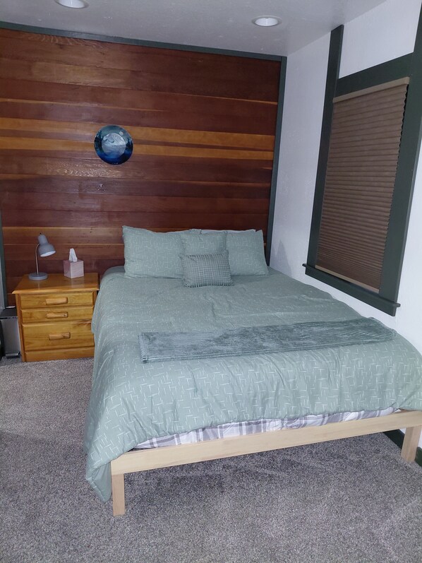 A bedroom with a wooden headboard, a bed with patterned bedding, a bedside table with a lamp, and a wall-mounted shelf.