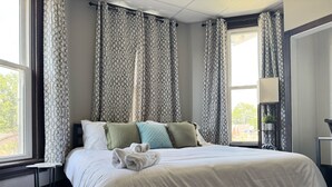 A neatly made bed with pillows and a folded towel, a bedside table, and a window with curtains.