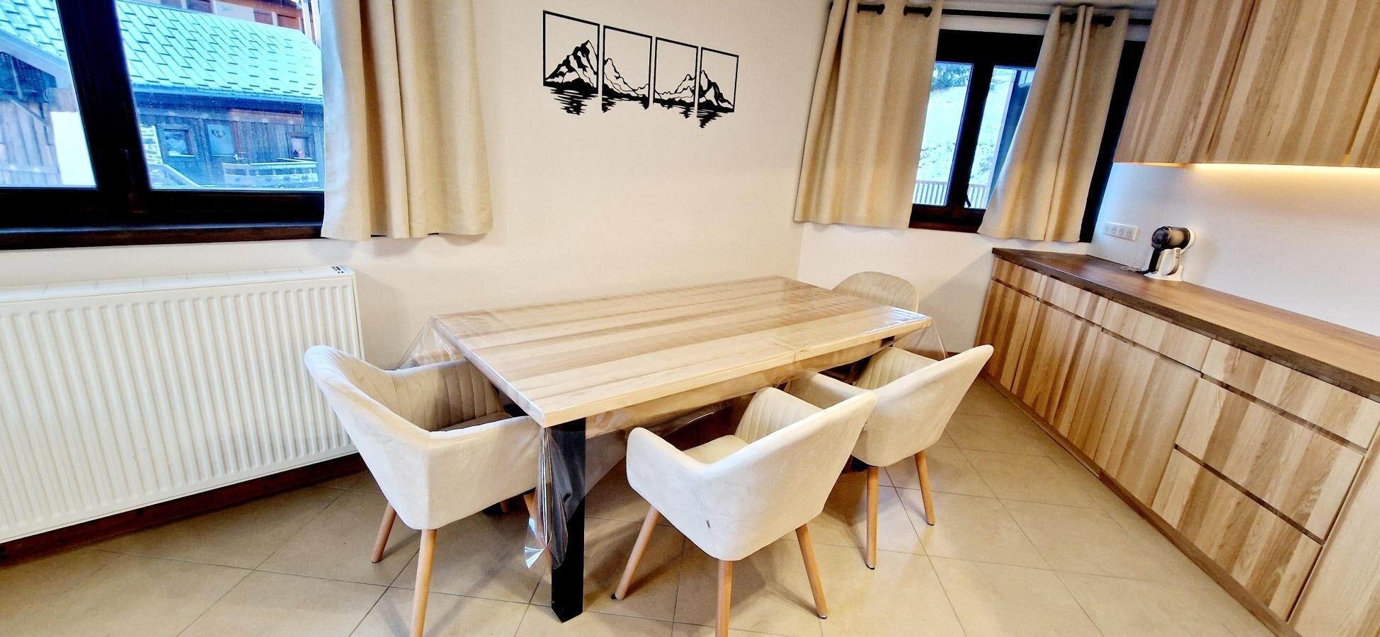 A modern kitchen with a wooden table and white chairs, a window with curtains, and a wall-mounted artwork.