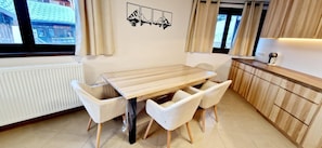 A modern kitchen with a wooden table and white chairs, a window with curtains, and a wall-mounted artwork.