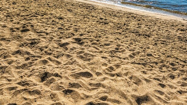 Ein Strand mit klarem, blauem Wasser, sandigem Ufer und einer bewaldeten Hügelkette im Hintergrund.
