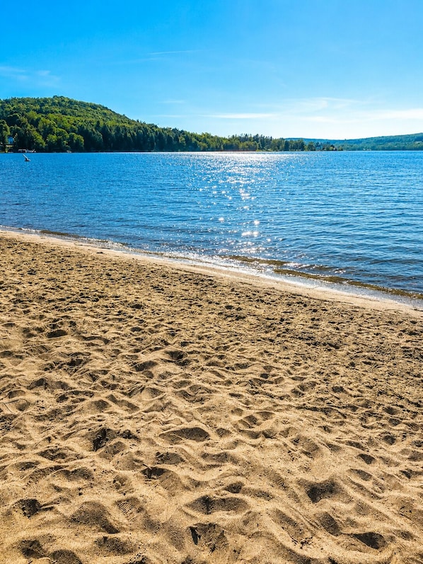 A beach with clear blue water, sandy shore, and a forested hill in the background.