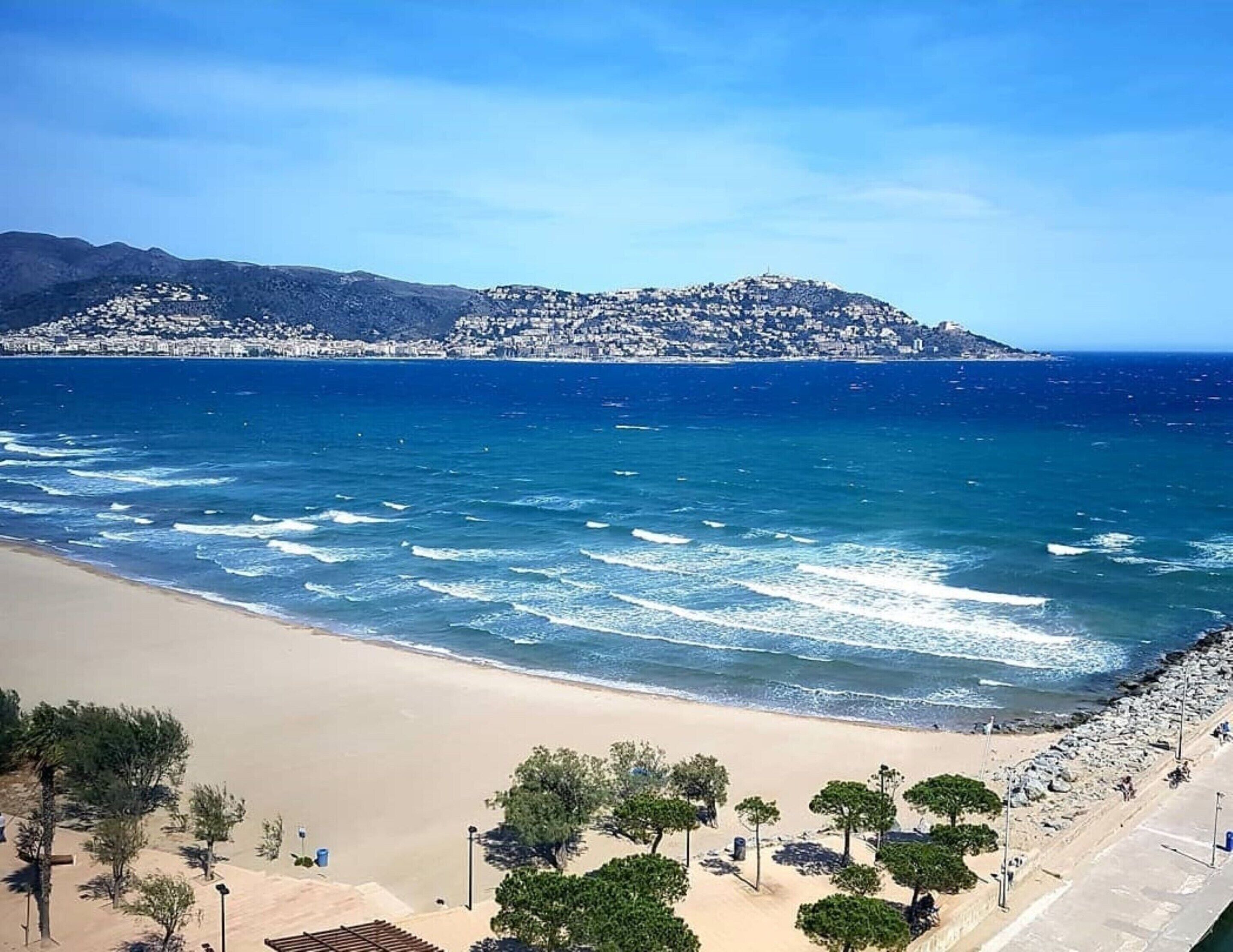 Ein Küstenstrand mit sandigem Ufer, einem klaren blauen Meer und einer Stadt auf einem Hügel im Hintergrund.