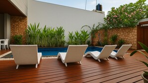 Un área junto a la piscina con tres sillones blancos de mimbre, una pared de piedra y abundante follaje.