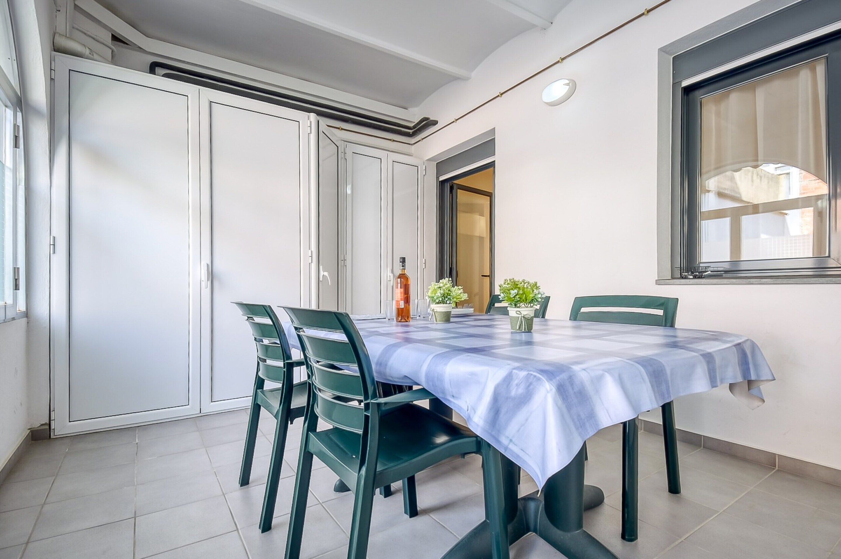 Zona pranzo con tavolo ricoperto da tovaglia blu, circondato da sedie verdi. Sulla tavola ci sono piante in vaso e una bottiglia di vino.