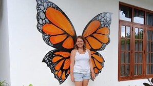Una persona de pie frente a una pared con un gran mural de mariposa. Hay plantas en macetas frente a la pared.