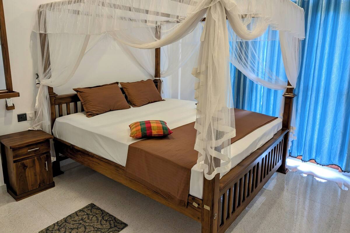 Una cama con dosel de madera con ropa de cama blanca y marrón, una mesita de noche pequeña y una almohada colorida.