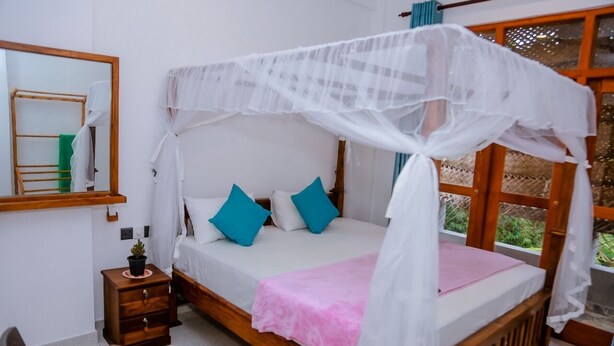 Una cama con dosel, una mesita de noche con una planta y una ventana con vistas a zonas verdes.