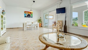 A spacious living area with a glass-top round table, white furniture, a large window with a view, and a TV mounted on the wall. - Apartment in Roses Near Canyelles Beach (Roses)