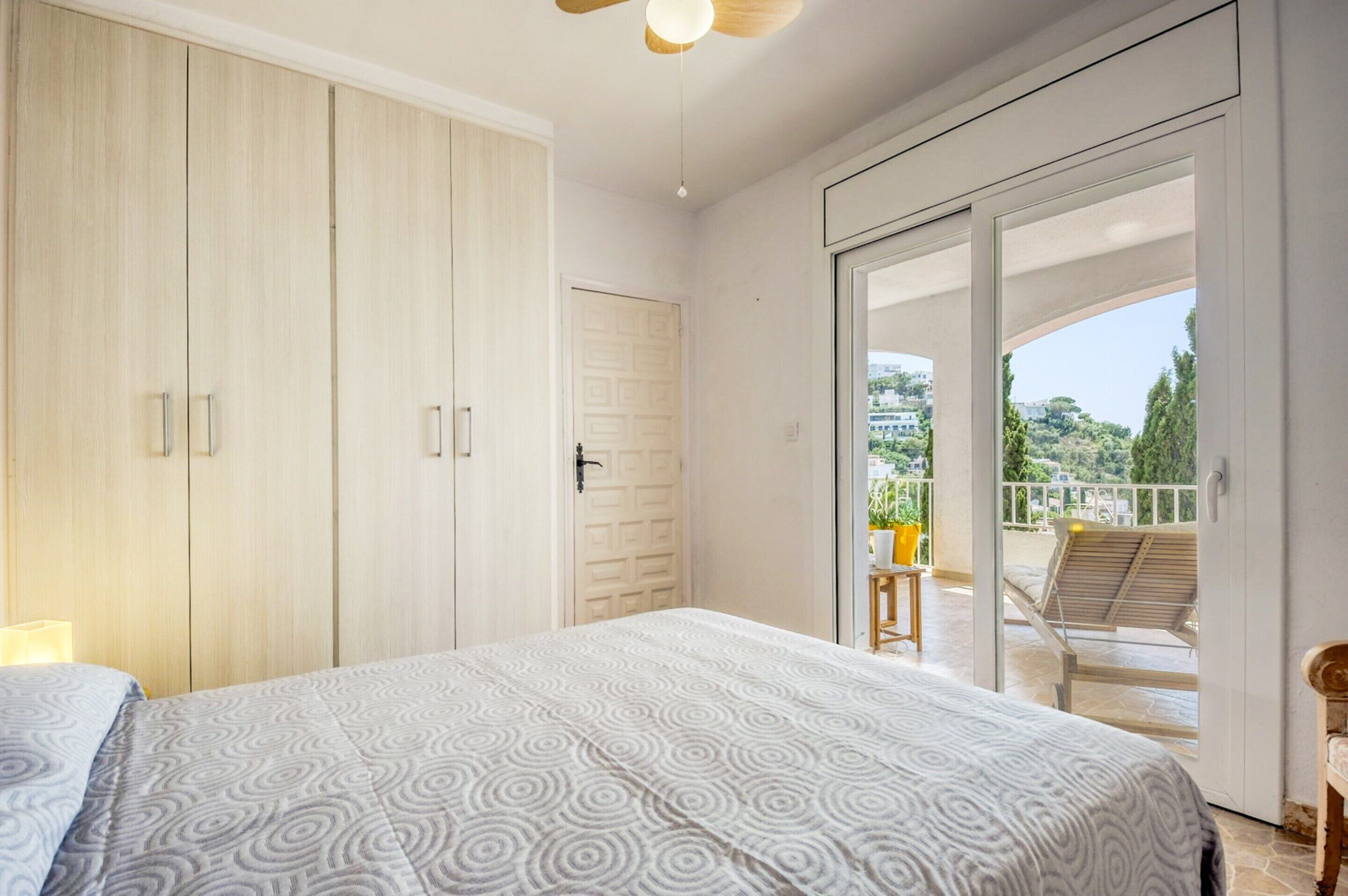 A bedroom with a bed, a wardrobe, and a sliding glass door leading to a balcony with a chair and a table.
