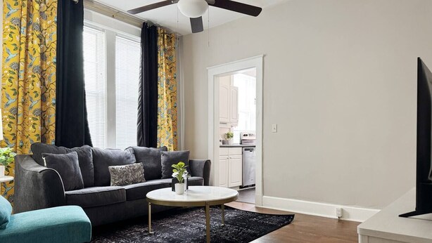 Un salon avec un canapé gris, un banc turquoise, une table basse ronde et un ventilateur de plafond.