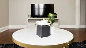Une table ronde avec un plateau en marbre blanc et des pieds dorés, un objet noir posé sur la table et une plante en pot à droite.