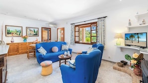 A living room with a blue sofa, a coffee table, and a TV. There are two windows with curtains, a kitchen area with wooden cabinets, and a dining table. - Can Pansa - Three Bedroom Villa, Sleeps 6 (Pollensa)
