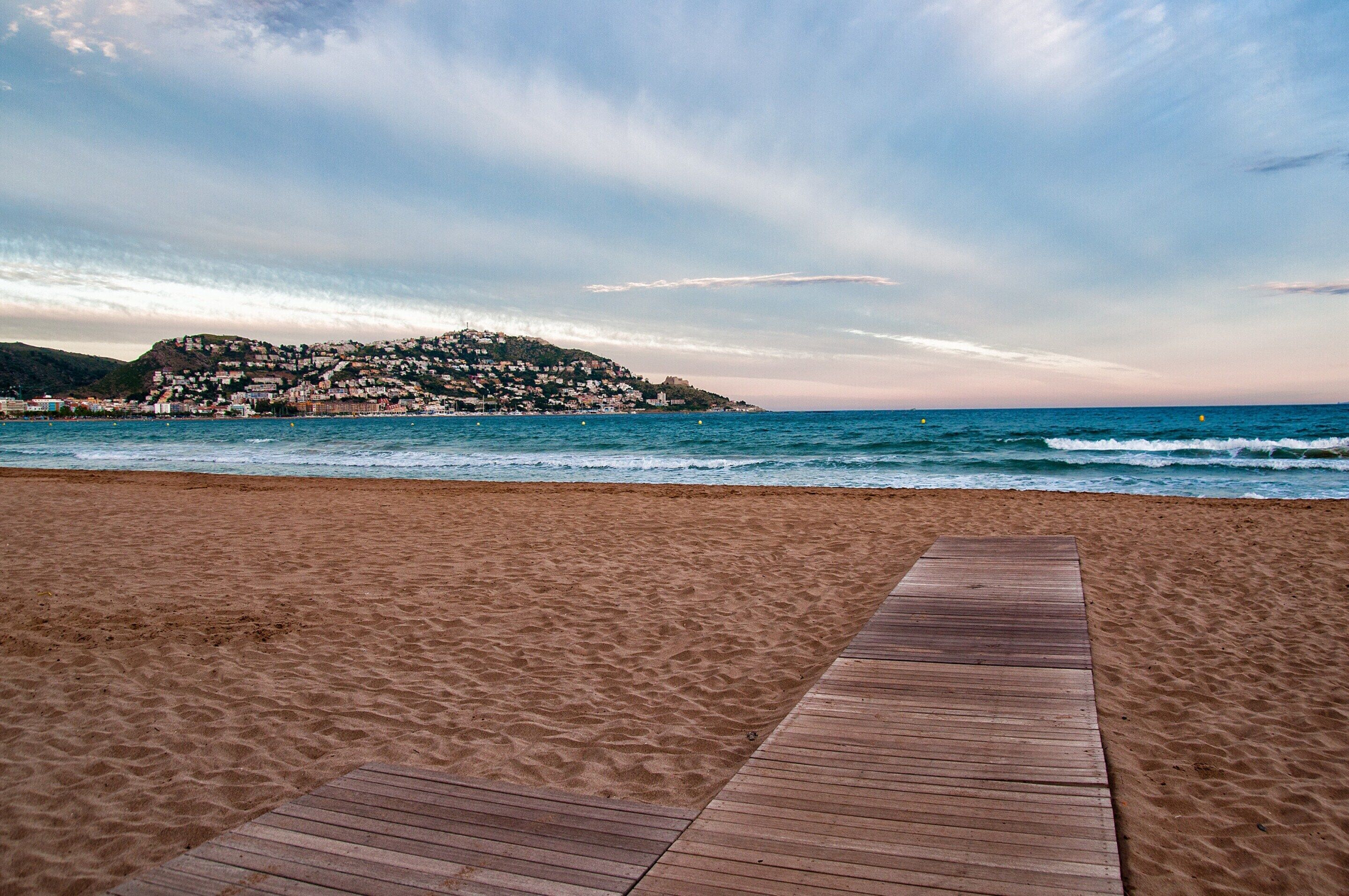 A sandy beach with a wooden walkway leading to the sea, overlooking a hillside with buildings.