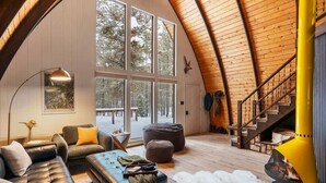 A cozy cabin interior with a large window, a fireplace, a staircase, and a leather sofa. - Retro A-Frame w/ Hot Tub, Perfect Couple's Getaway (Island Park)