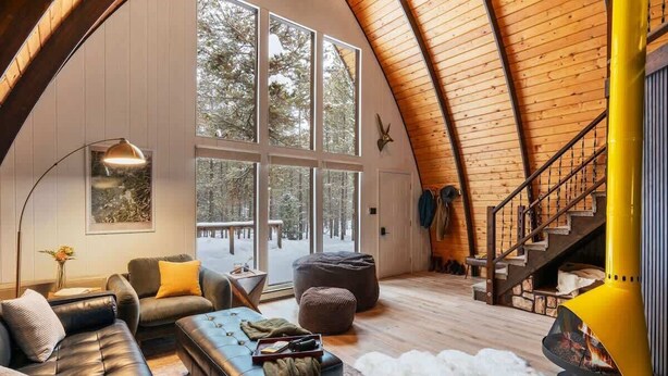 A cozy cabin interior with a large window, a fireplace, a staircase, and a leather sofa.