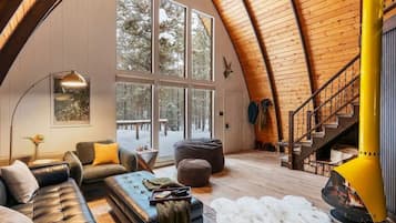 A cozy cabin interior with a large window, a fireplace, a staircase, and a leather sofa.