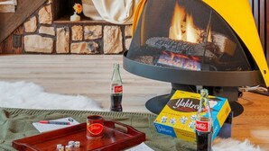 A cozy living room with a fireplace, a wooden staircase, and a dining area with a tray, dice, and a bottle of Coca-Cola. - Retro A-Frame w/ Hot Tub, Perfect Couple's Getaway (Island Park)