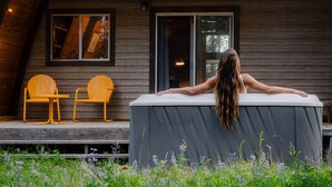 A two-story wooden cabin with a covered outdoor seating area and a large window overlooking the landscape.
