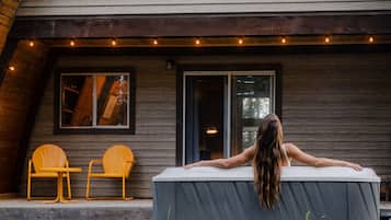 A two-story wooden cabin with a covered outdoor seating area and a large window overlooking the landscape.
