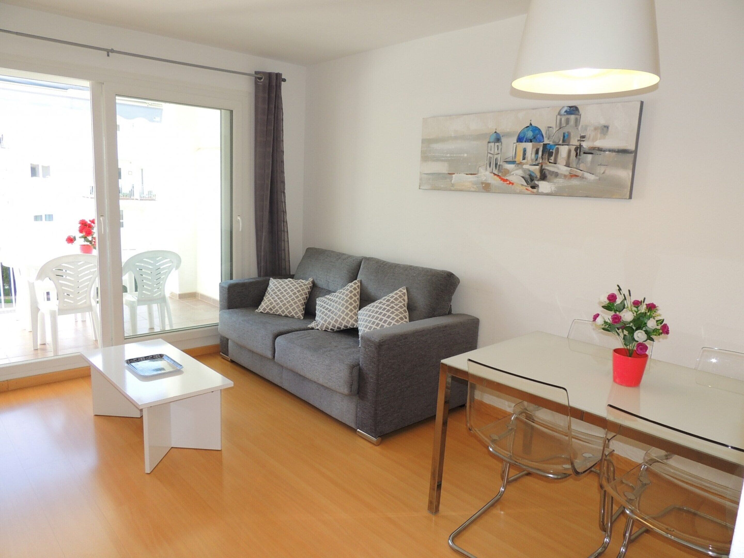 Una sala moderna con un sofá gris, una mesa de centro blanca y una mesa de comedor de vidrio con sillas. Hay un cuadro en la pared y una ventana con cortinas.