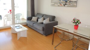 Una sala moderna con un sofá gris, una mesa de centro blanca y una mesa de comedor de vidrio con sillas. Hay un cuadro en la pared y una ventana con cortinas.