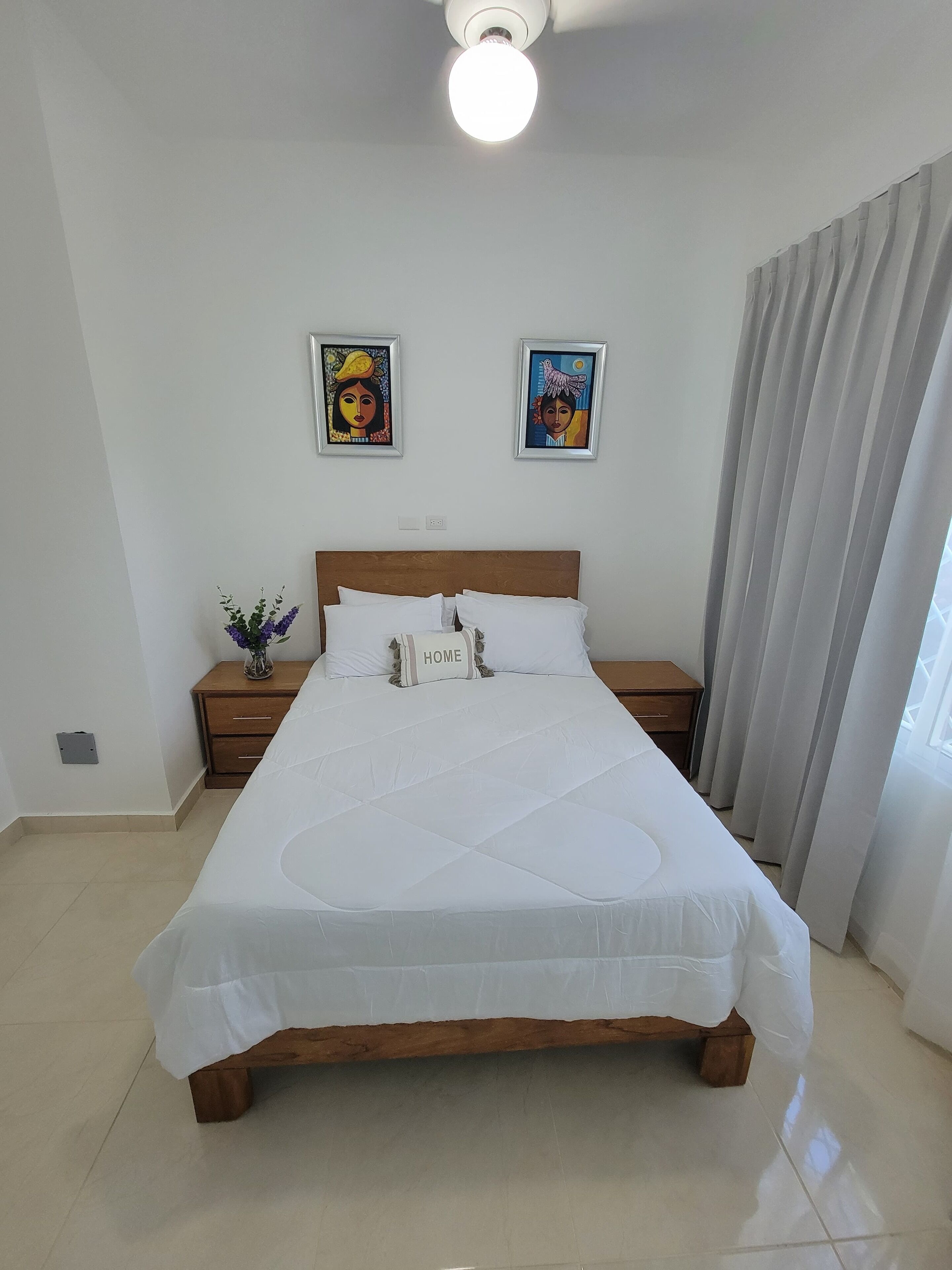 Una cama bien hecha con cabecera de madera, una mesita de noche con un florero y dos cuadros enmarcados en la pared.