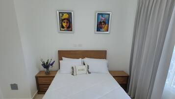 Una cama bien hecha con cabecera de madera, una mesita de noche con un florero y dos cuadros enmarcados en la pared.
