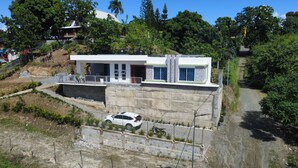 Una casa moderna de dos pisos con balcón, entrada para vehículos y un paisaje circundante con árboles y una colina.